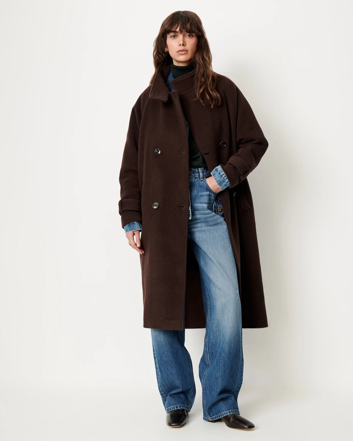 A model wearing a long, dark brown double-breasted coat over a dark green turtleneck sweater, blue jeans, and black shoes, standing against a plain off-white studio background.