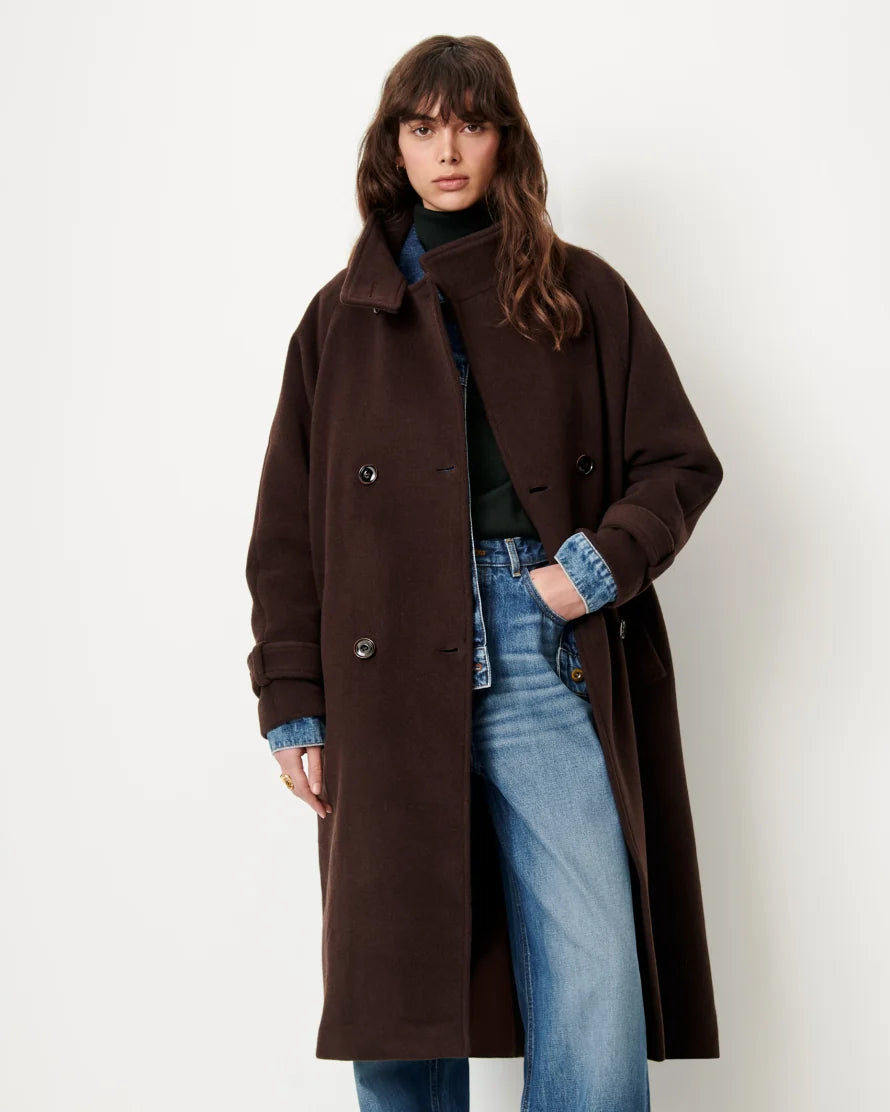 A woman modeling a long, dark brown double-breasted coat over a black top and blue jeans, standing against a plain off-white studio background.