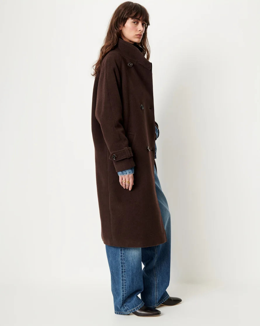 A model wearing a long, dark brown double-breasted coat over a blue denim shirt and wide-leg blue jeans, standing against a plain off-white studio background.