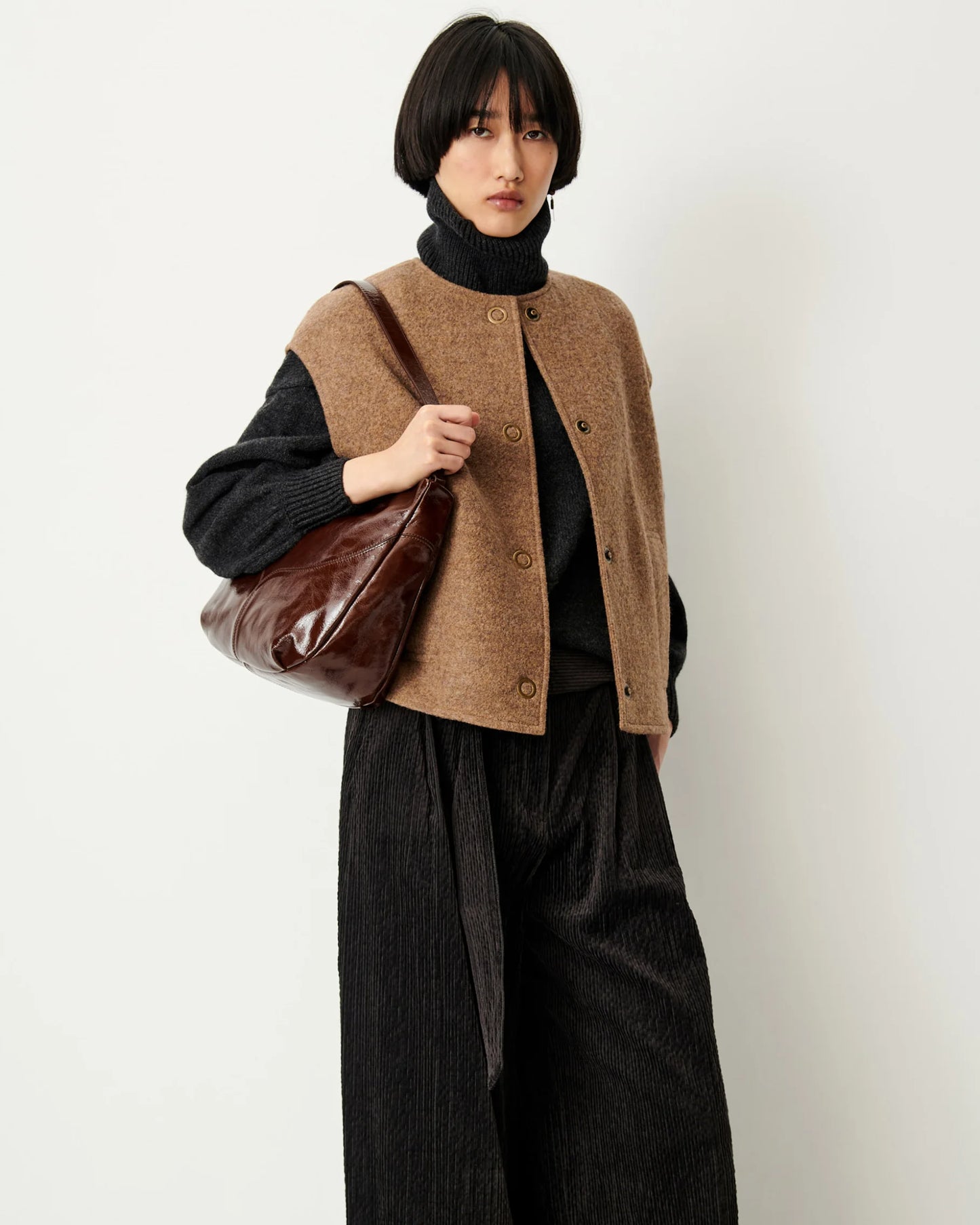 A model wearing a brown button-up knit vest over a black turtleneck sweater, paired with black wide-leg corduroy trousers and carrying a brown leather shoulder bag, set against a plain off-white background.