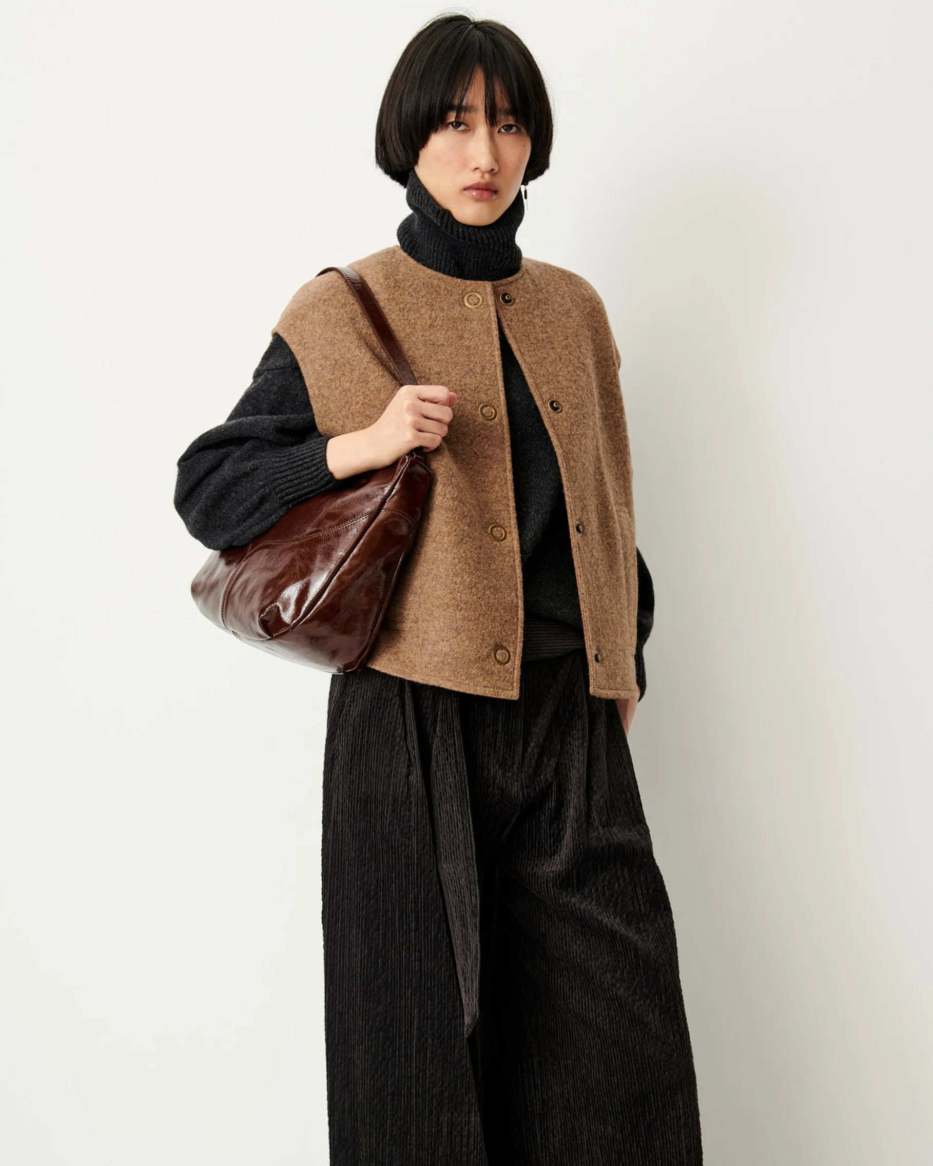 A model wearing a brown button-up knit vest over a black turtleneck sweater, paired with black wide-leg corduroy trousers and carrying a brown leather shoulder bag, set against a plain off-white background.