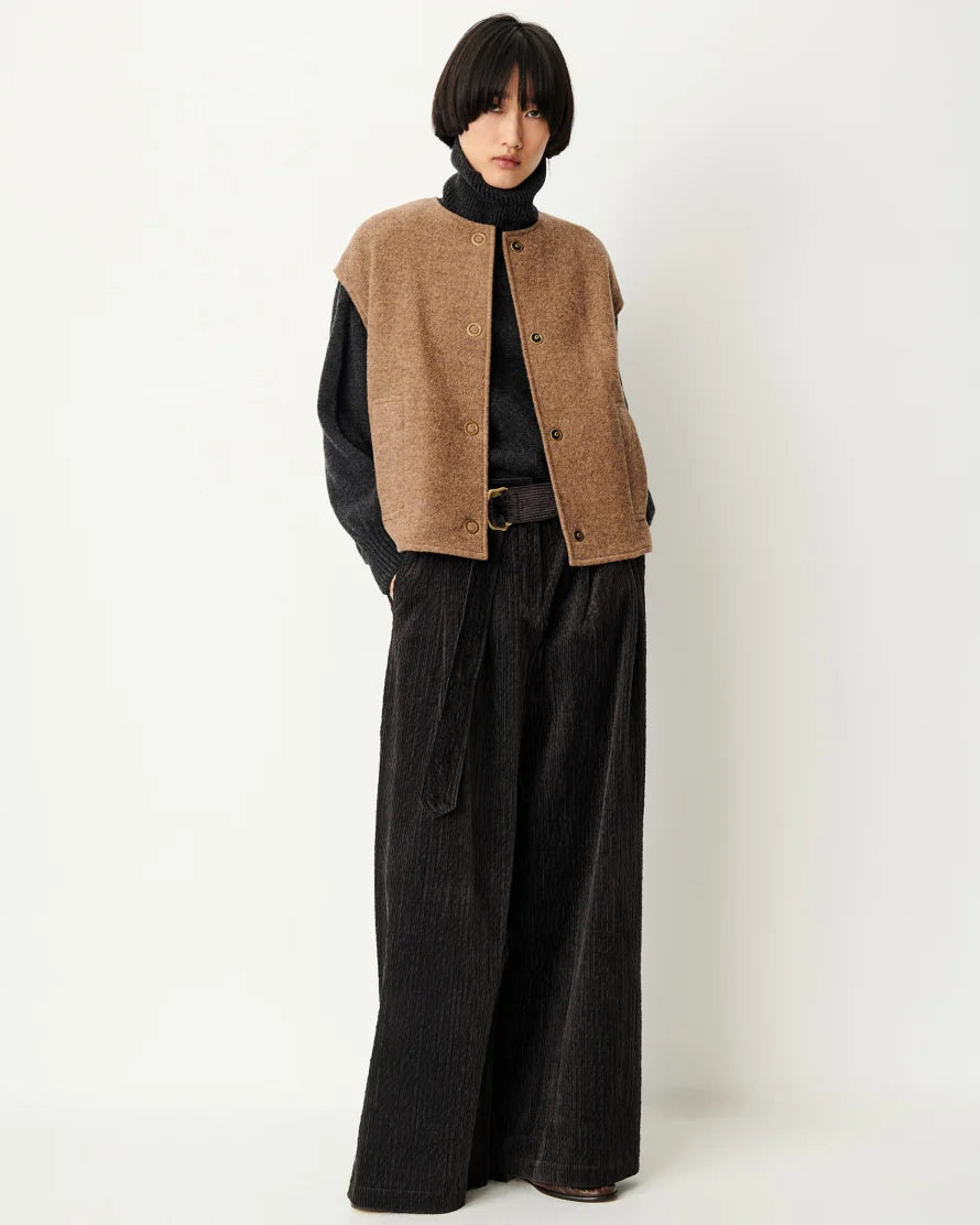 A model stands against a plain off-white studio background wearing a brown cropped button-front vest over a black turtleneck, paired with wide-leg black corduroy trousers.