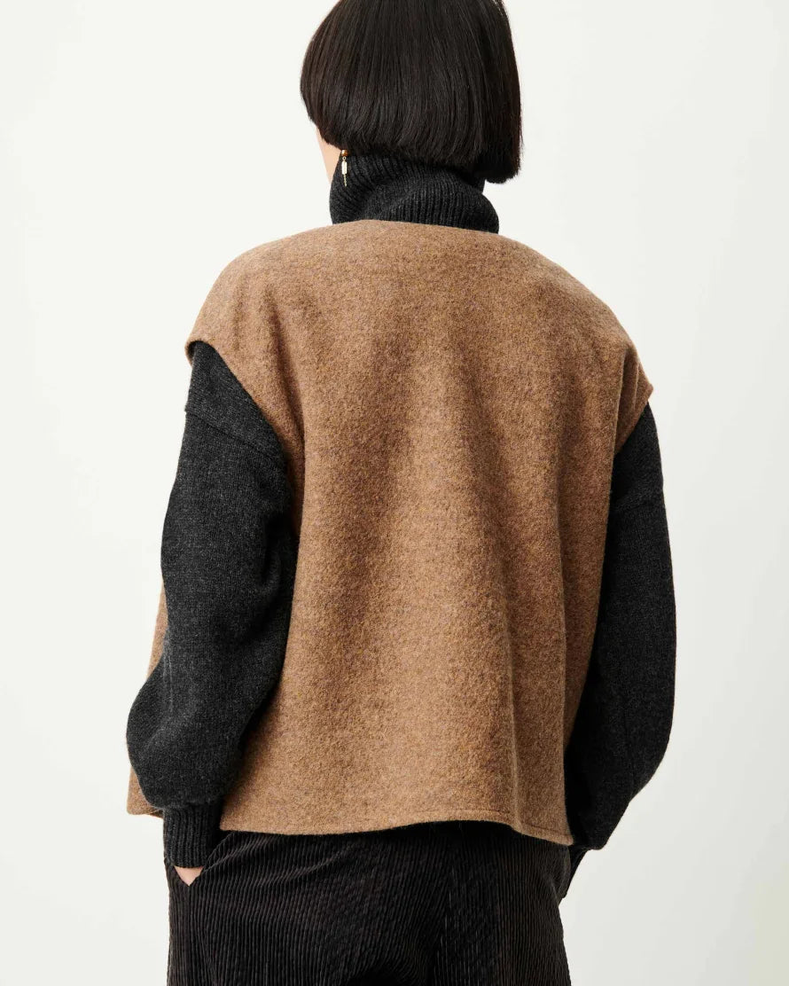 A person shown from the back wearing a cropped knit sweater vest in camel brown with dark charcoal sleeves, layered over a black turtleneck and dark ribbed pants, against a plain light background.