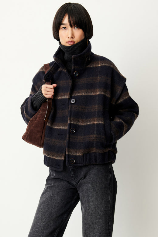 A model wearing a dark navy and brown plaid button-up jacket with a high ribbed turtleneck collar, paired with dark gray jeans and a brown shoulder bag, against a plain light gray background.