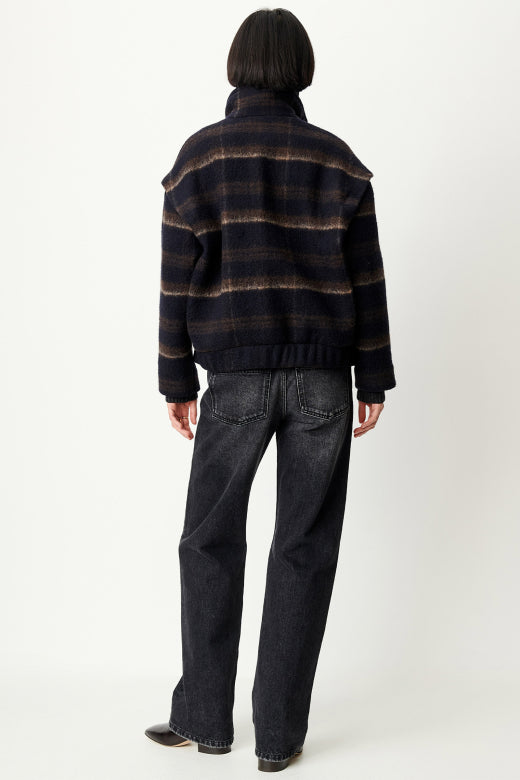 Rear view of a model wearing a dark navy and brown plaid button-up jacket with a high ribbed turtleneck collar, paired with dark gray jeans and a brown shoulder bag, against a plain light gray background