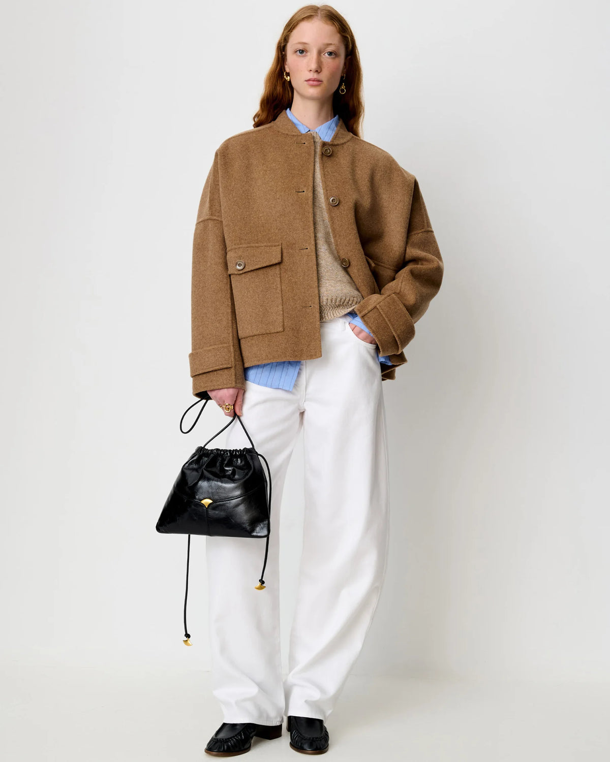 A model stands against a light gray studio background wearing a camel brown cropped jacket over a beige sweater and blue striped shirt, paired with white wide-leg trousers, black shoes, and holding a small black drawstring handbag.