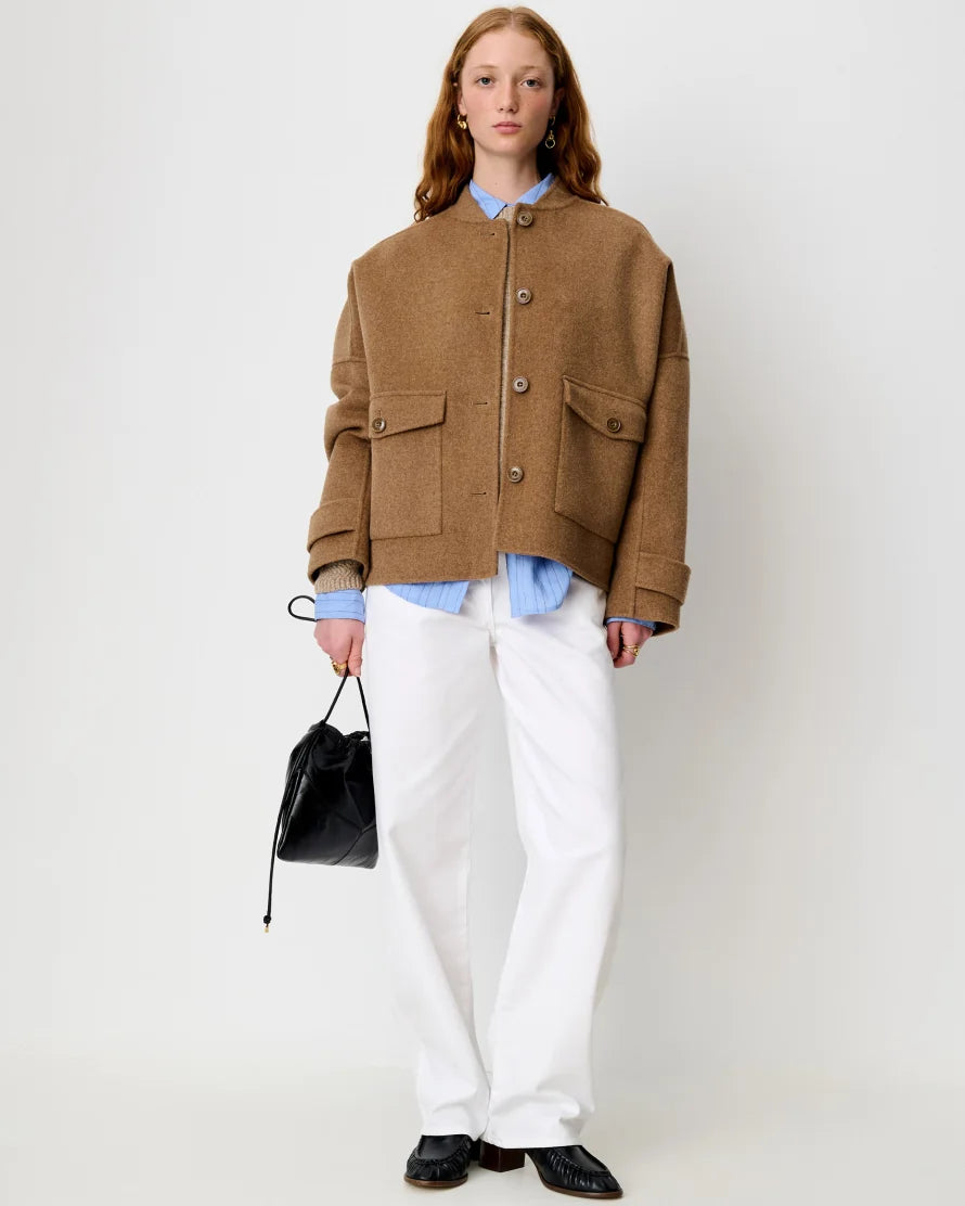 A model shown wearing a light brown jacket over a light blue shirt. She is wearing white pants and standing against a white background.