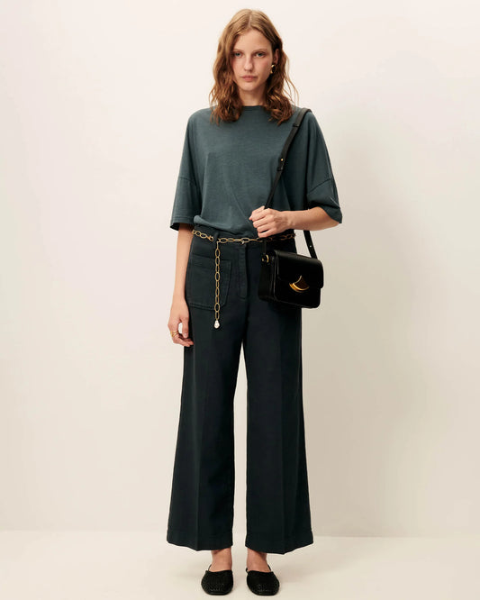 Woman wearing a green top and green pants with a black handbag and gold chain belt on a white background