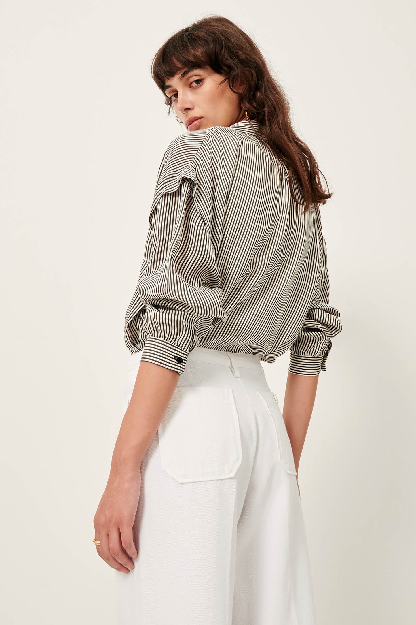 Picture, full length of a young woman with shoulder length wavy brown hair and brown eyes wearing a stripped shirt and white pants. facing away from camera looking over here shoulder at camera