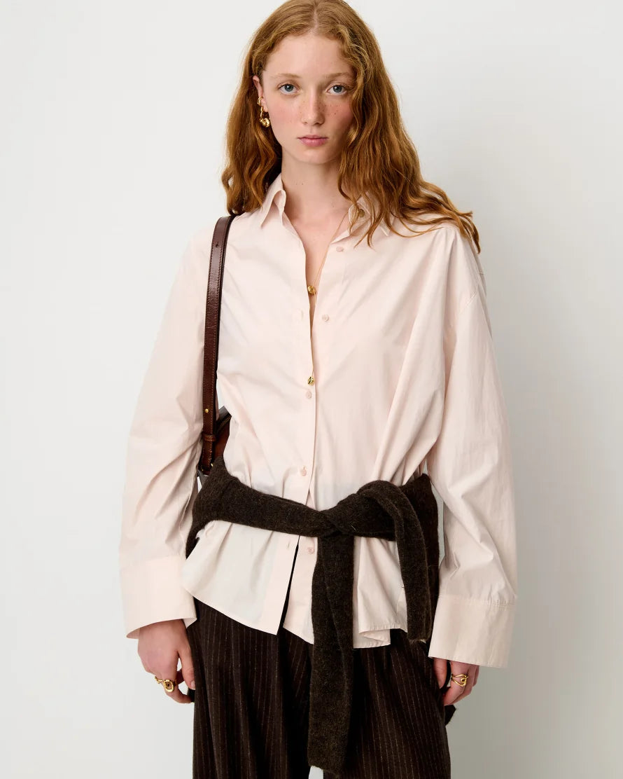 A model with red hair is wearing a pale pink button-down shirt tied at the waist with a dark brown fuzzy belt, paired with dark brown corduroy trousers and a brown shoulder bag, against a plain light gray background.