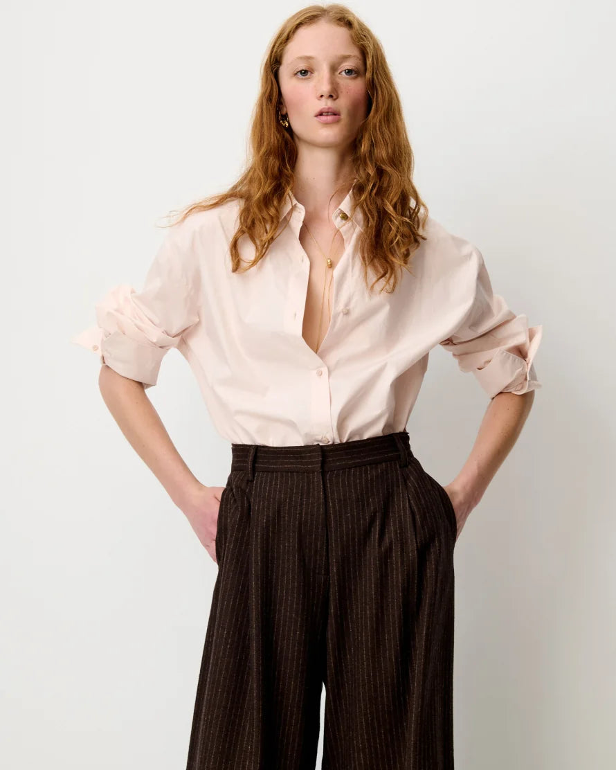 A model with long wavy red hair is wearing a pale pink button-down shirt with ruffled sleeves and dark brown pinstriped wide-leg trousers, standing against a light gray studio background.