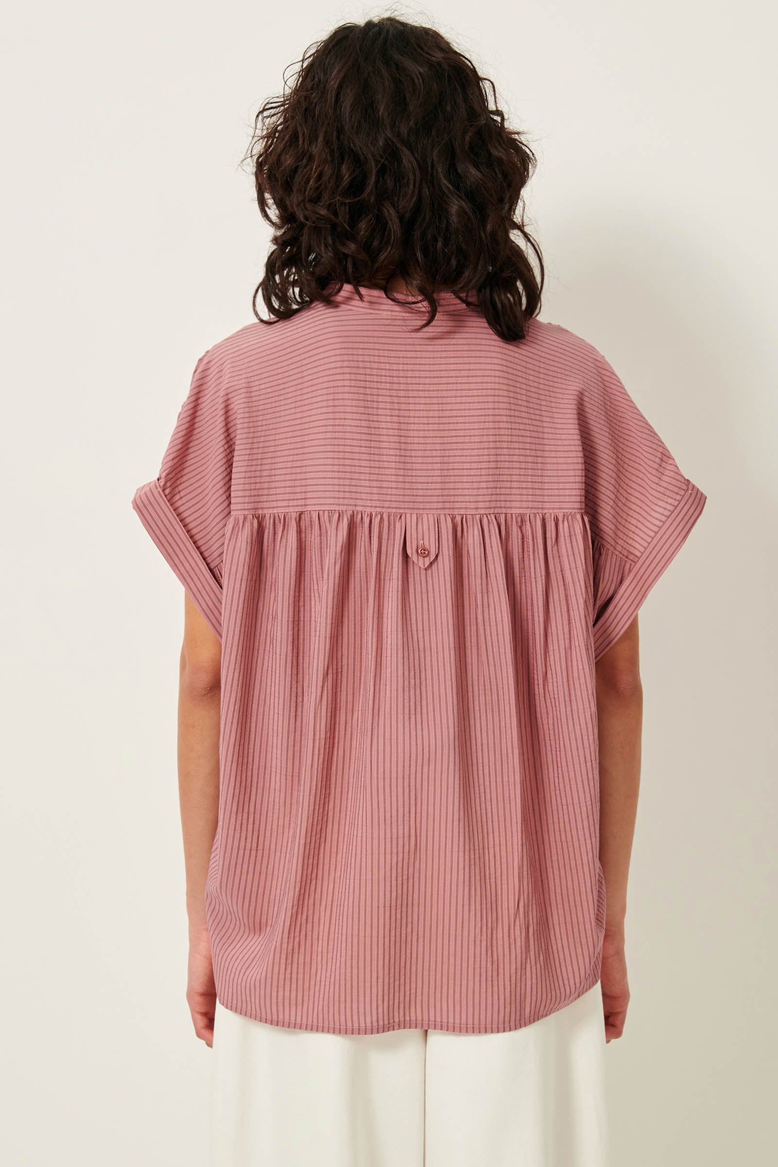 Picture of  the back of young women from the waist up with black wavy shoulder length hair dark brown eyes wearing a striped rose color sleeveless shirt.