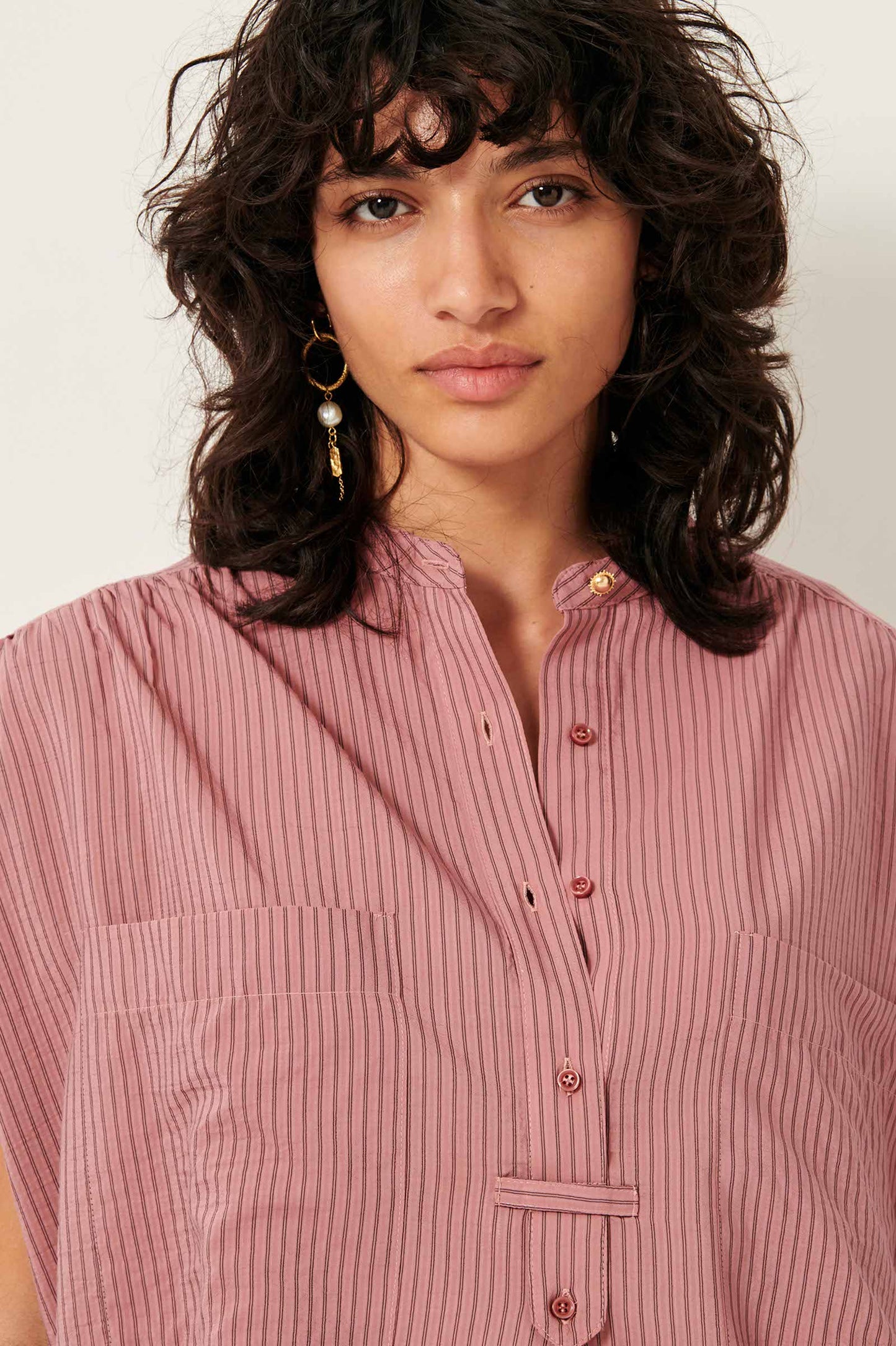 Picture close up of young women from the waist up with black wavy shoulder length hair dark brown eyes wearing a striped rose color sleeveless shirt.