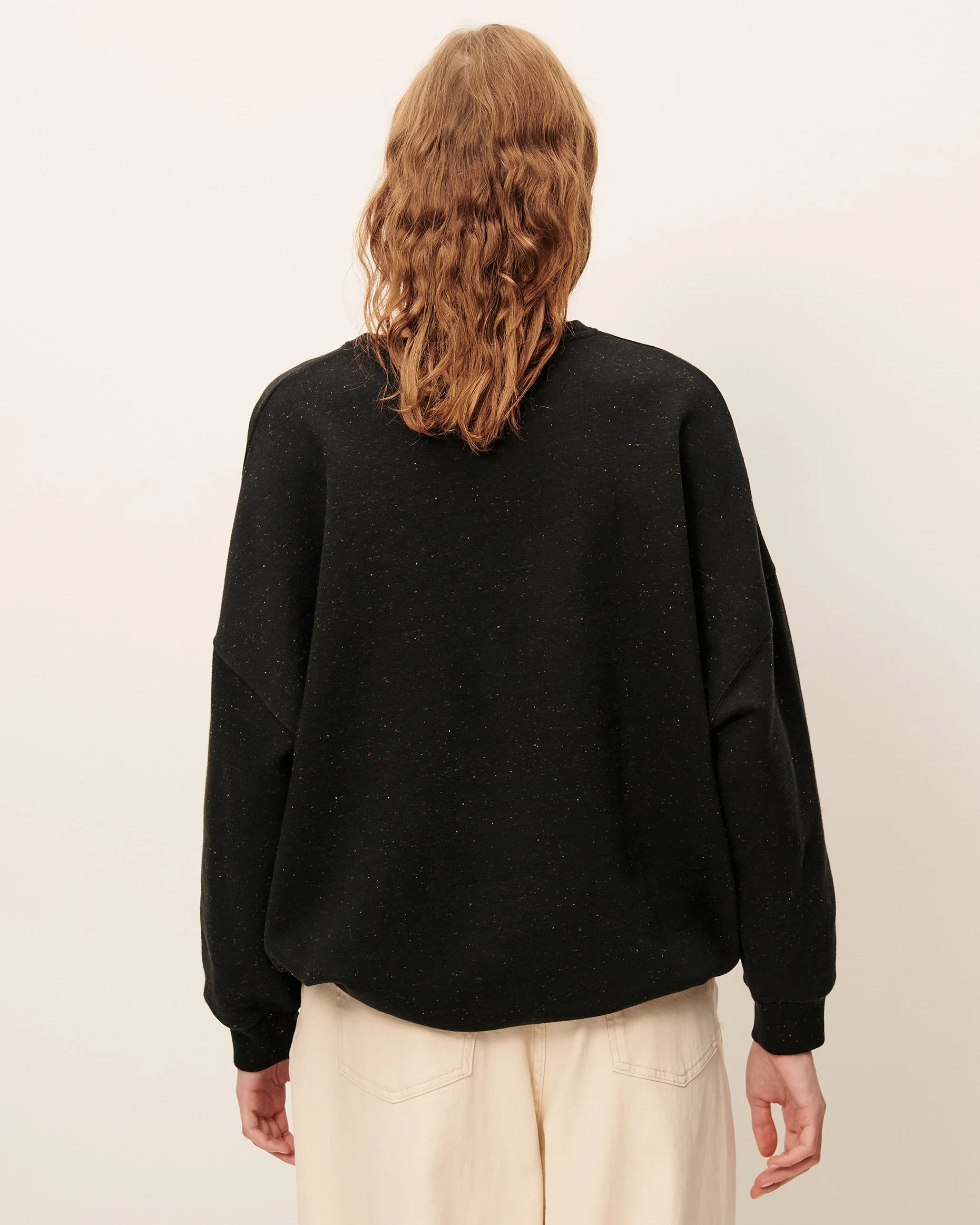 A person shown from the back wearing a loose black long-sleeve knit top and light beige pants against a plain off-white studio background.