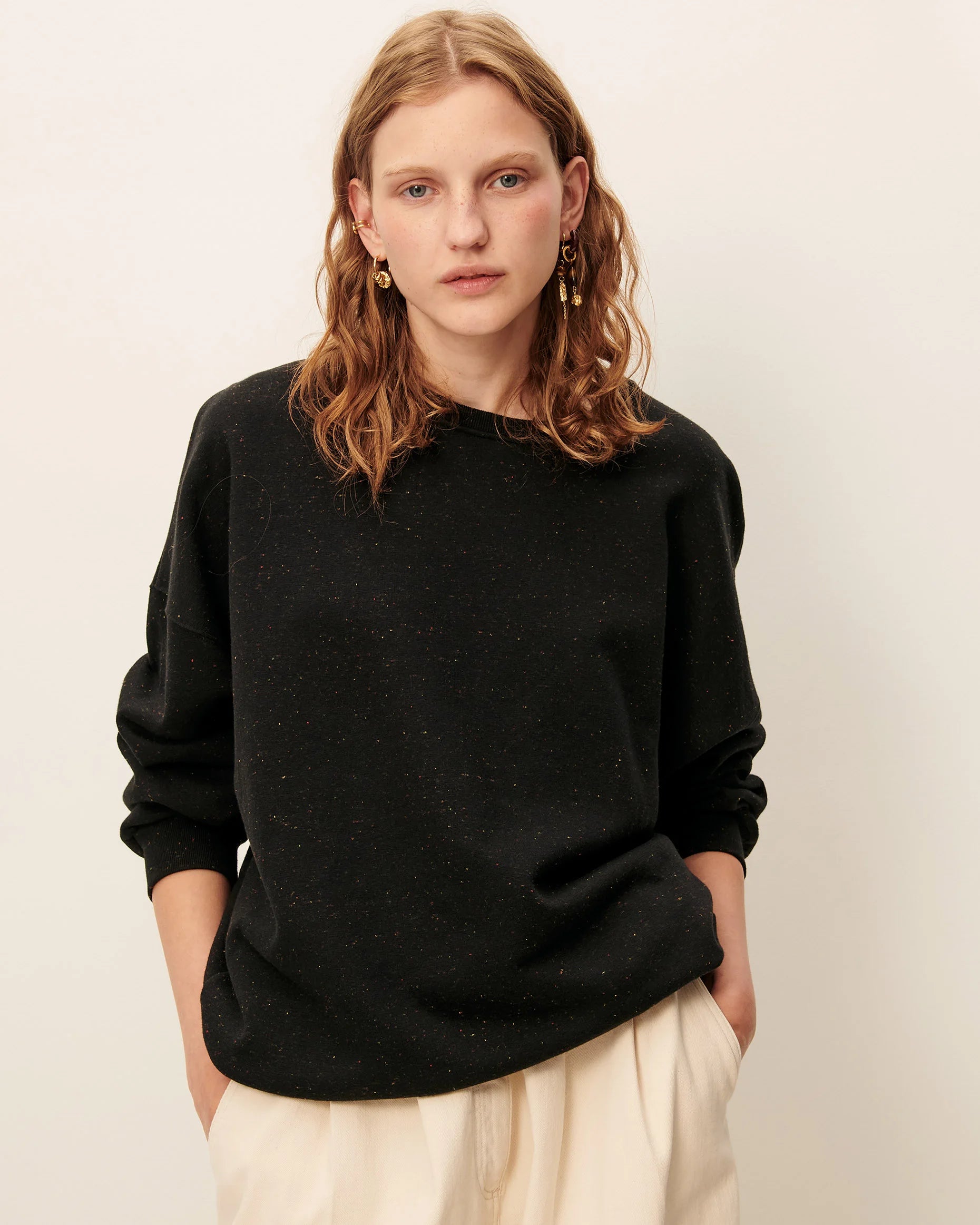 woman with blond hair wearing black sweatshirt and cream pants against white background