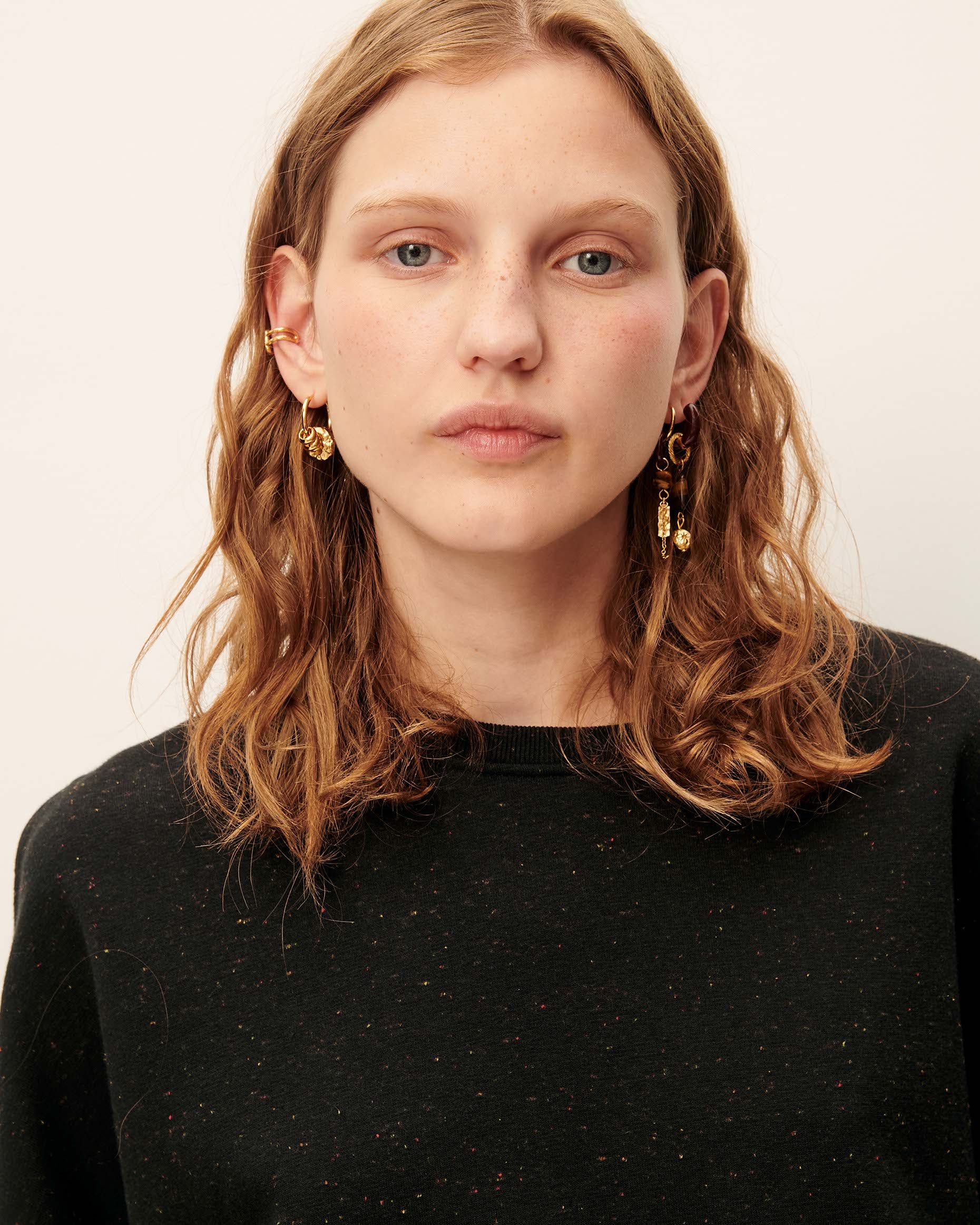 A close-up portrait of a woman with wavy light brown hair wearing gold dangle earrings and a black speckled knit top against a plain off-white background.