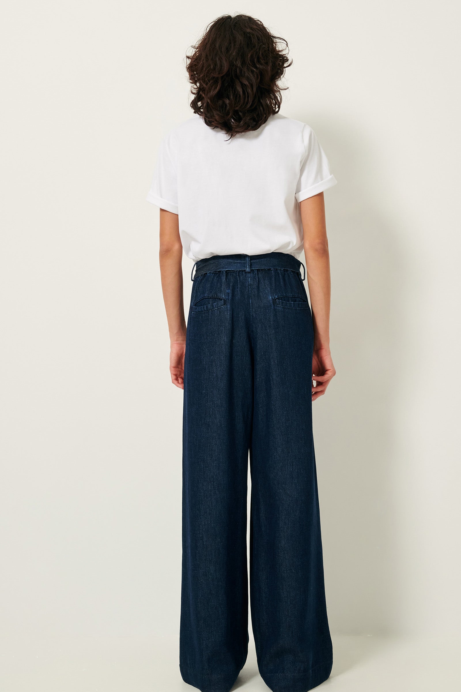 Picture of the back of Brown wavy haired young woman with brown eyes wearing a white shirt blue pants. 