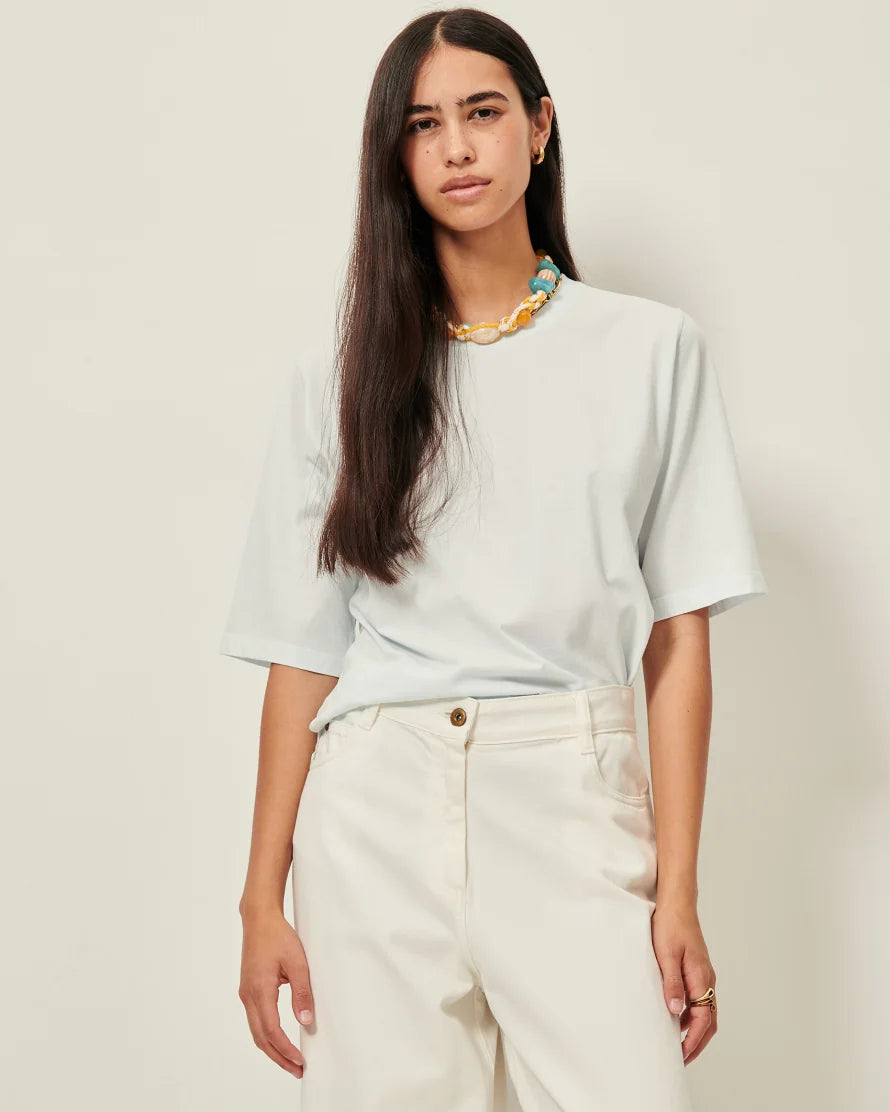 Model wearing an ice blue T-shirt with white jeans and a yellow necklace. White background