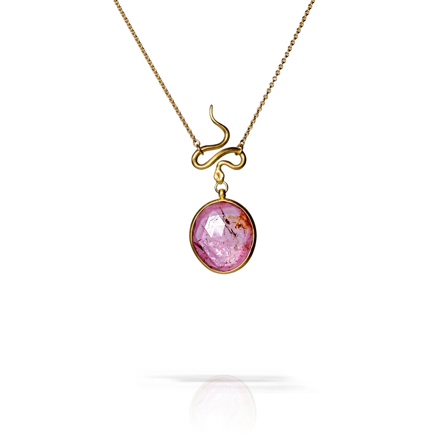 A gold pendant necklace featuring a circular faceted purple gemstone with inclusions, suspended from a curved gold chain, shown against a white background with a soft reflection below.