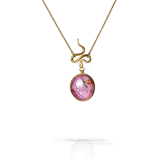 A gold pendant necklace featuring a circular faceted purple gemstone with inclusions, suspended from a curved gold chain, shown against a white background with a soft reflection below.