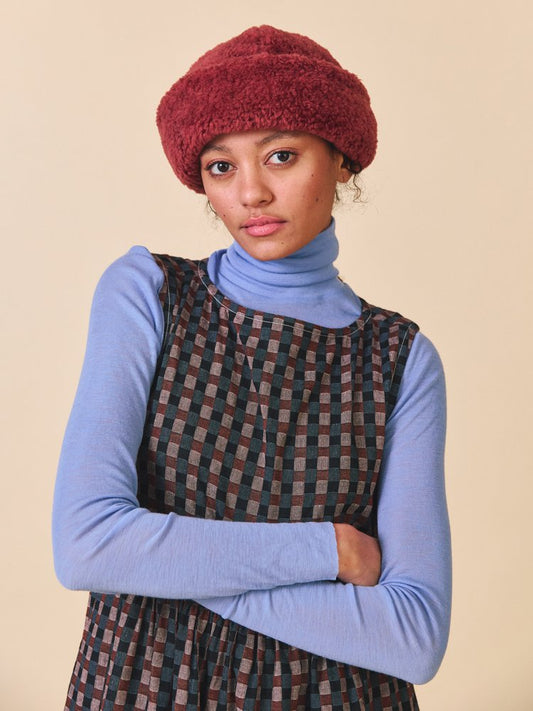 Woman wearing a red knitted hat, blue turtleneck, and plaid dress against a beige background