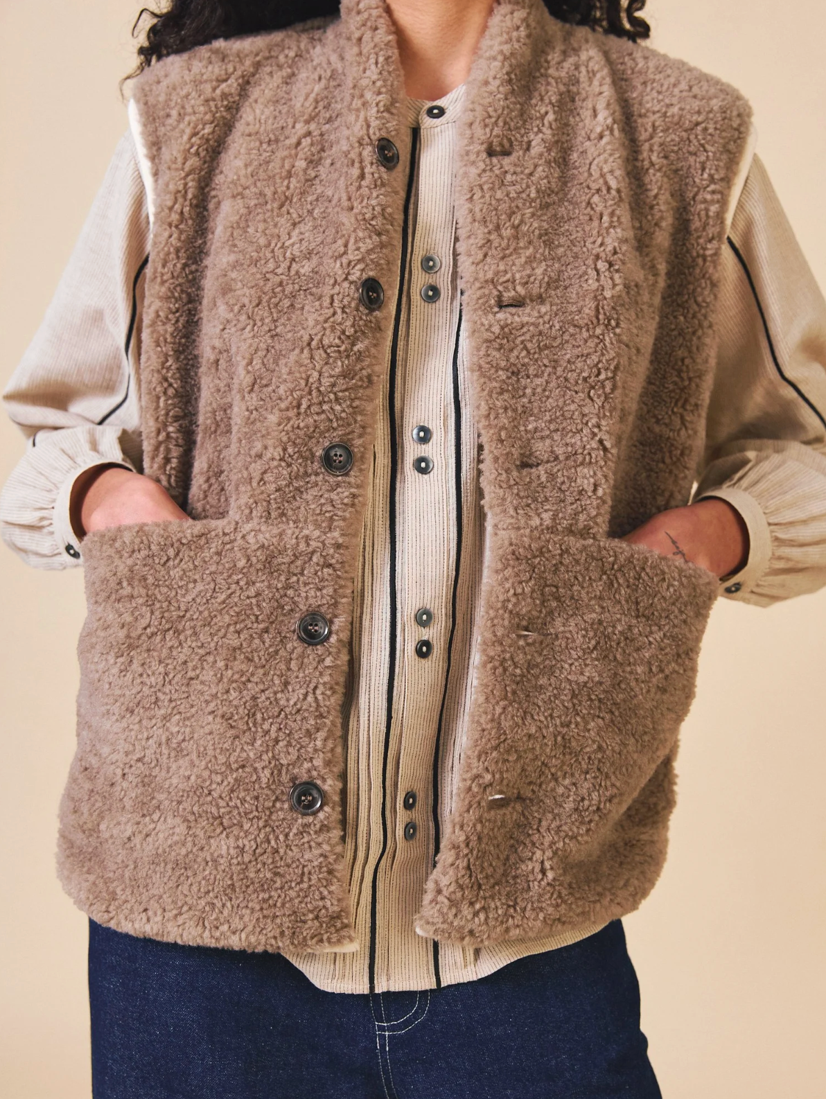 Person wearing a brown sherpa vest over a cream shirt with a beige background