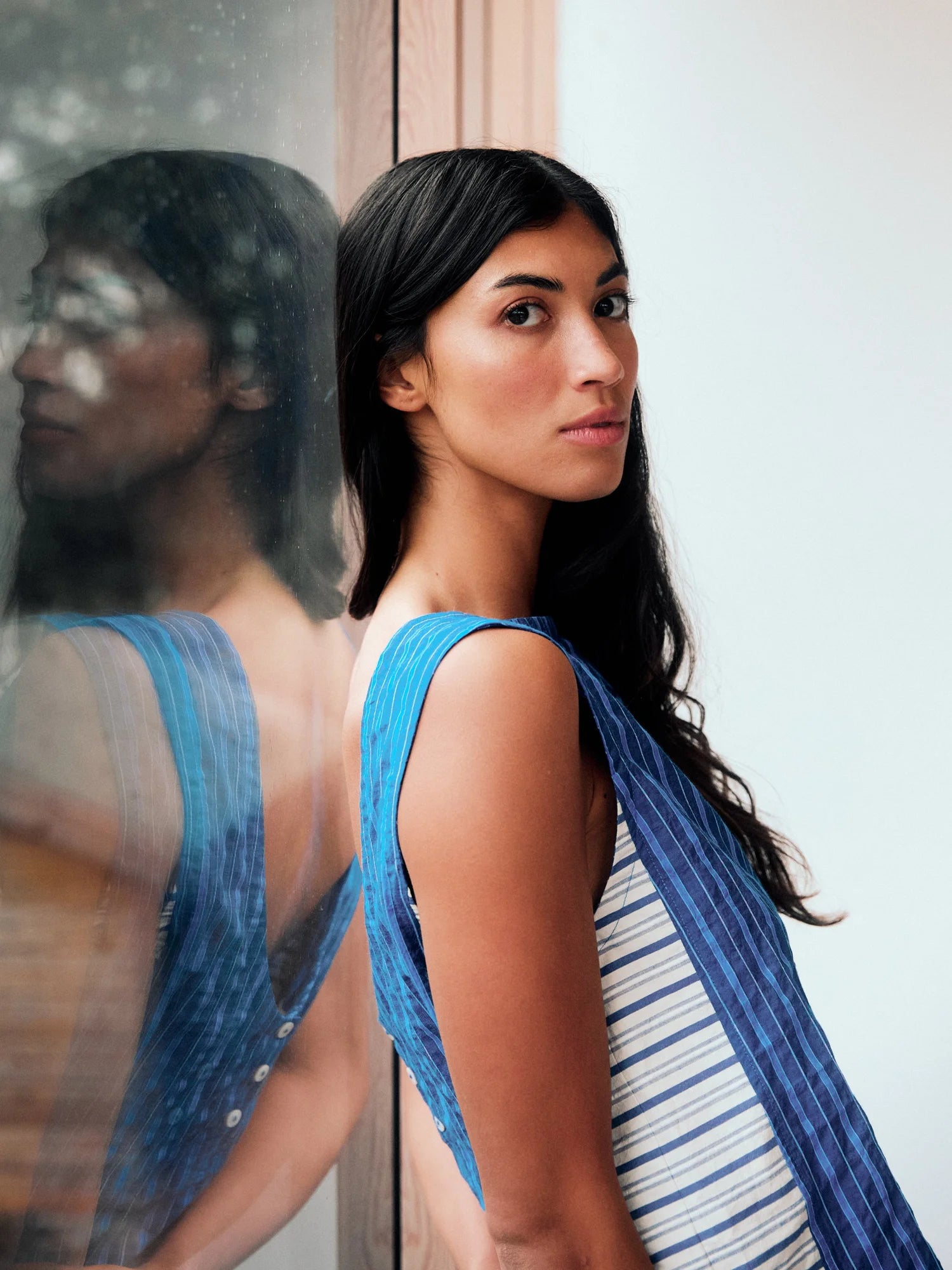 Woman wearing a stripe sleeveless top with a contrasting side panel. The model is leaning against a window with a reflection of her and the back view of the top 