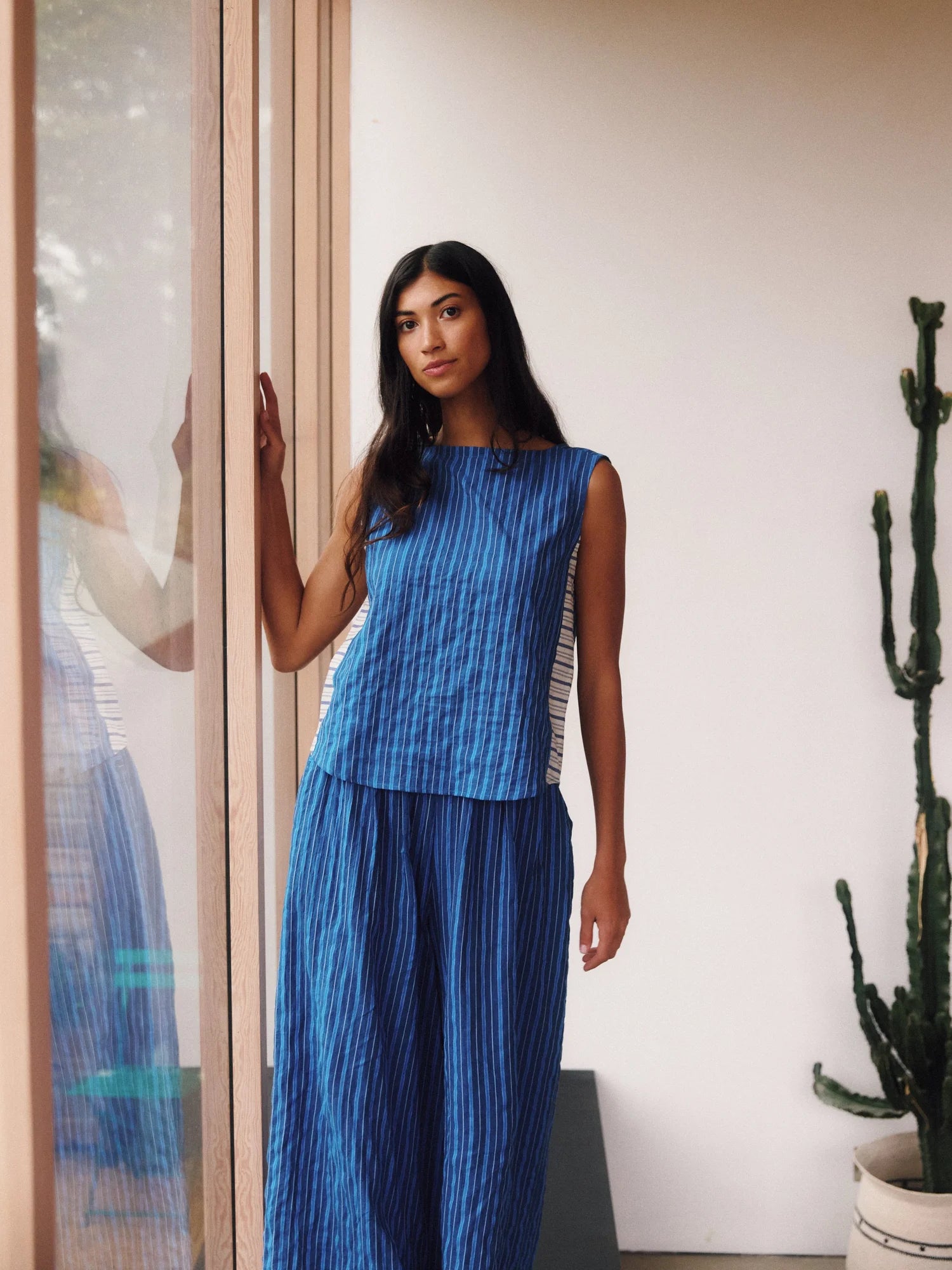 Woman wearing a blue stripe sleeveless top and pant outfit standing indoors with a plant in the background