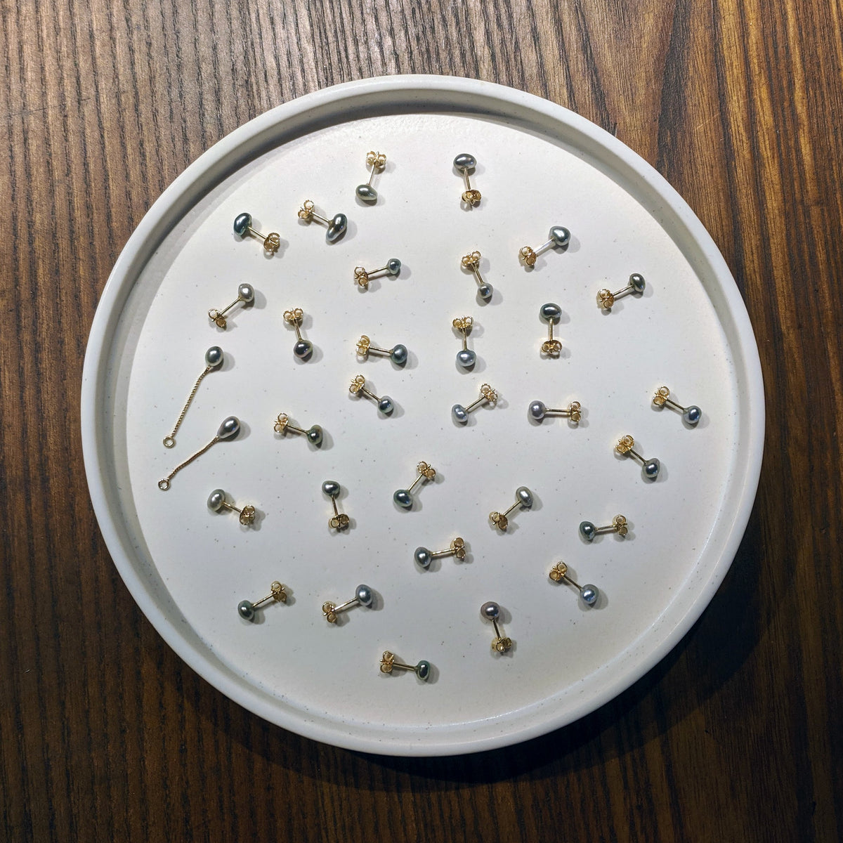 A white circular tray filled with multiple small pearl stud earrings, photographed from above on a dark wood surface.
