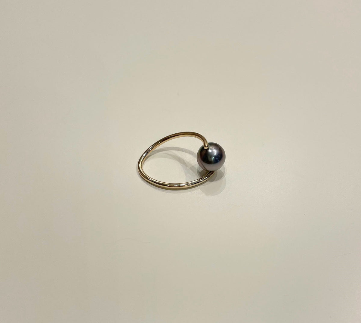A minimalist gold ring featuring a single dark gray pearl bead, photographed on a clean, light beige background.