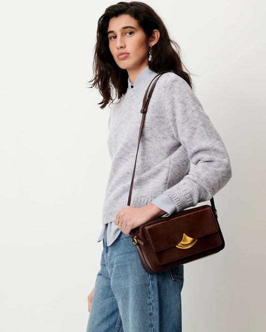A woman wearing a light gray knit sweater and blue jeans carries a brown leather shoulder bag with a gold-tone turn-lock clasp against a plain off-white background.