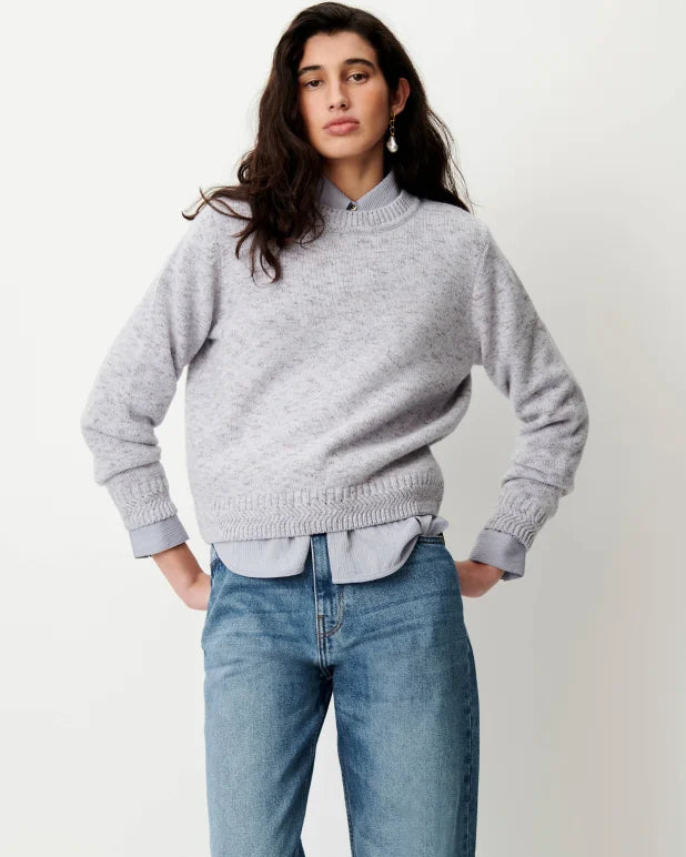 A woman modeling a light gray knit mock-neck sweater over a light blue collared shirt and blue jeans, standing against a plain off-white studio background.