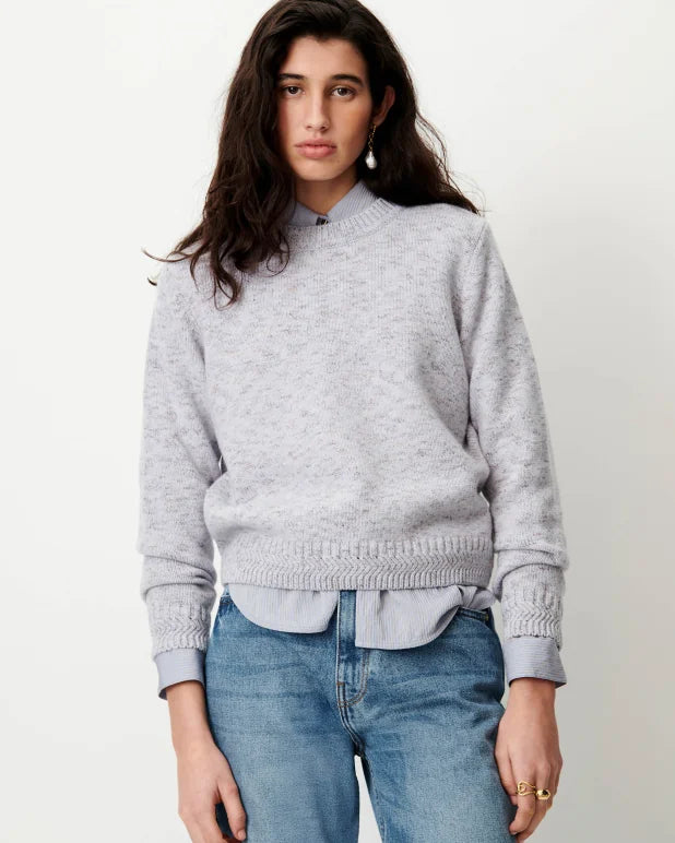 A woman wearing a light gray knit mock neck sweater over a white collared shirt and blue jeans, standing against a plain off-white background.