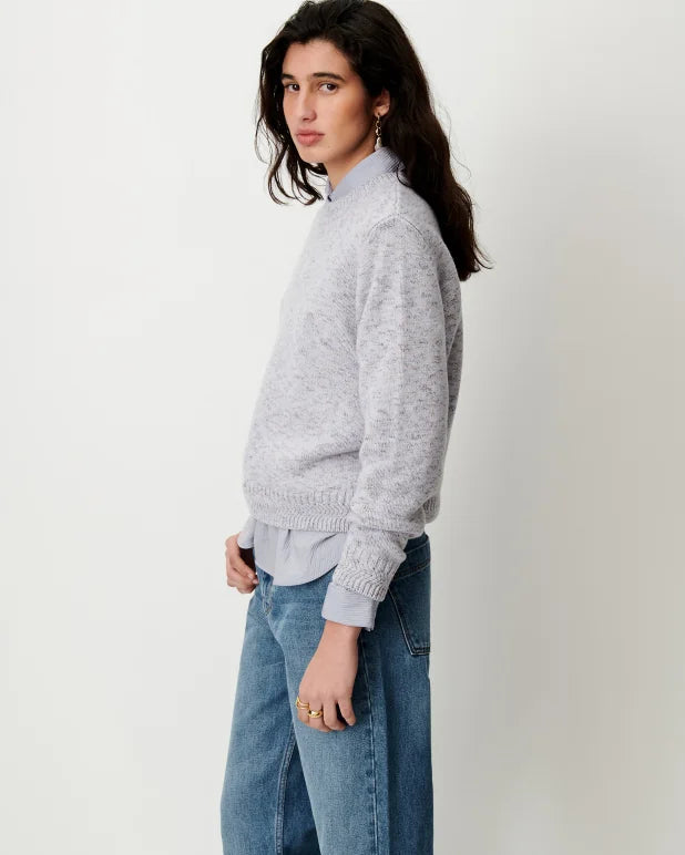 A woman wearing a light gray knit mock-neck sweater over a light blue collared shirt and blue jeans, standing against a plain off-white studio background.