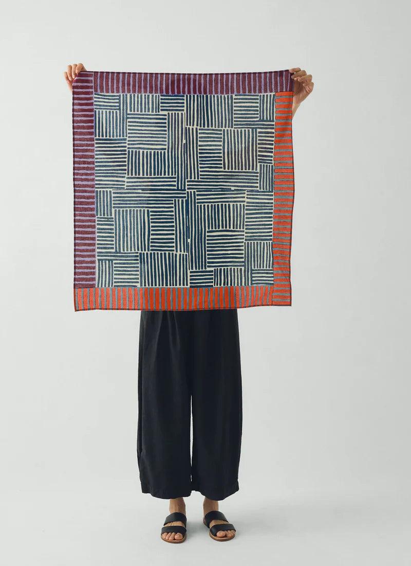A model holding up a block print bandana in front of her face and torso to show the full size and pattern. White background