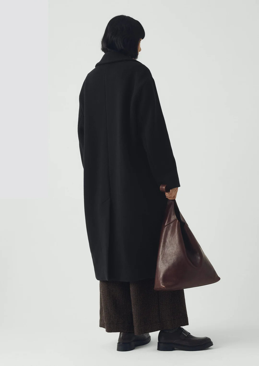 Person wearing a long black coat holding a brown bag on a white background