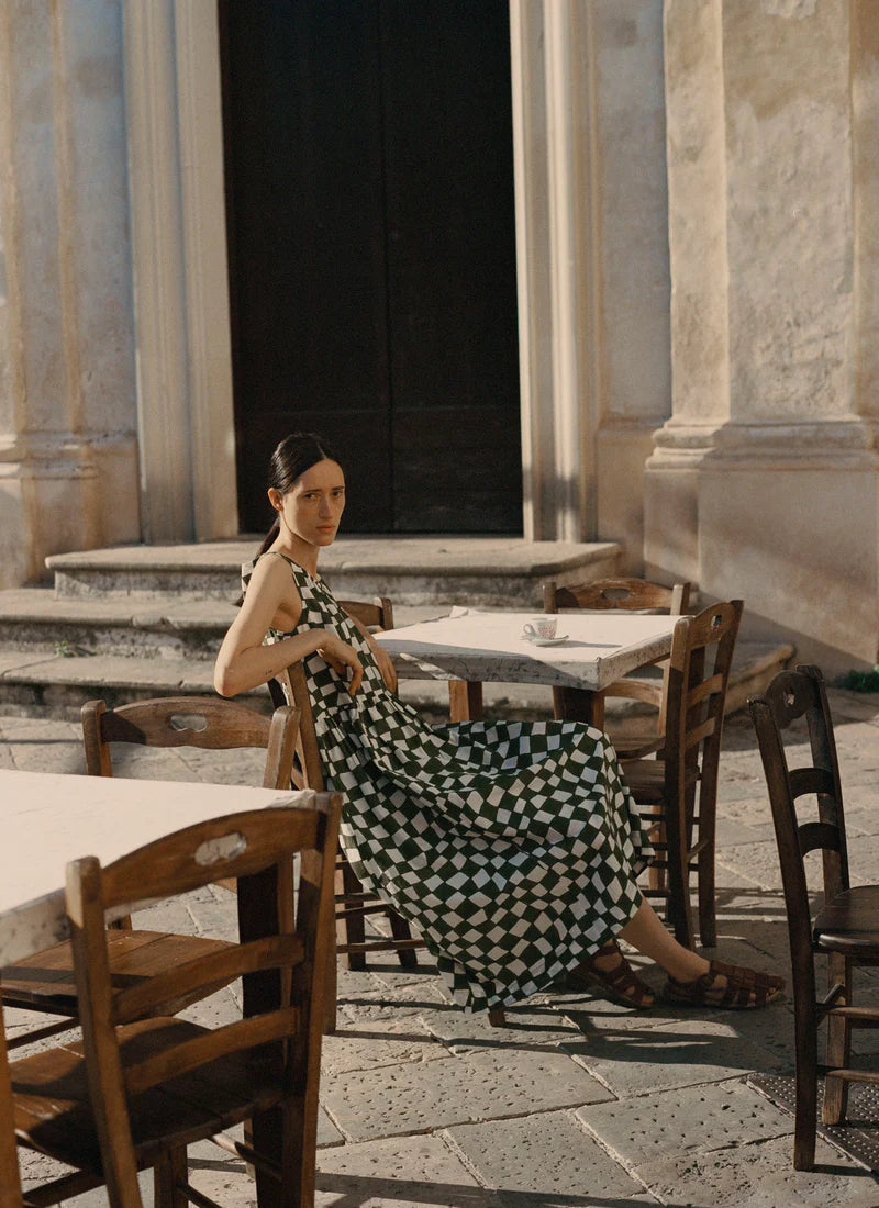 Woman in a checkered dress sitting at an outdoor cafe with classical architecture in the background