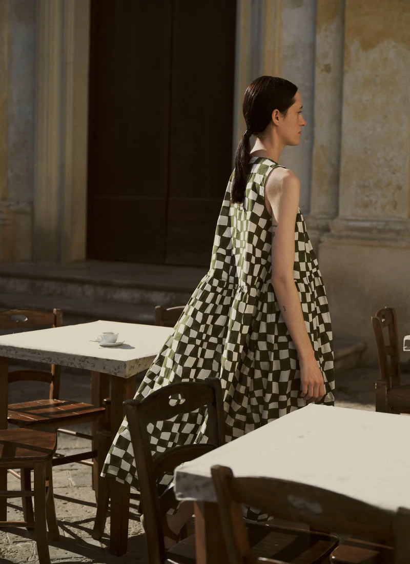 Woman in a checkered dress standing in an old, rustic room with tables and chairs.