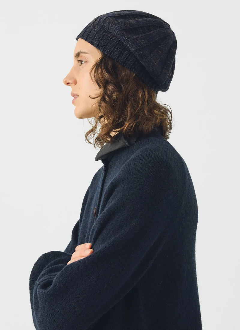 A person in profile wearing a dark navy ribbed knit beanie and a matching dark navy knit sweater against a light gray studio background.