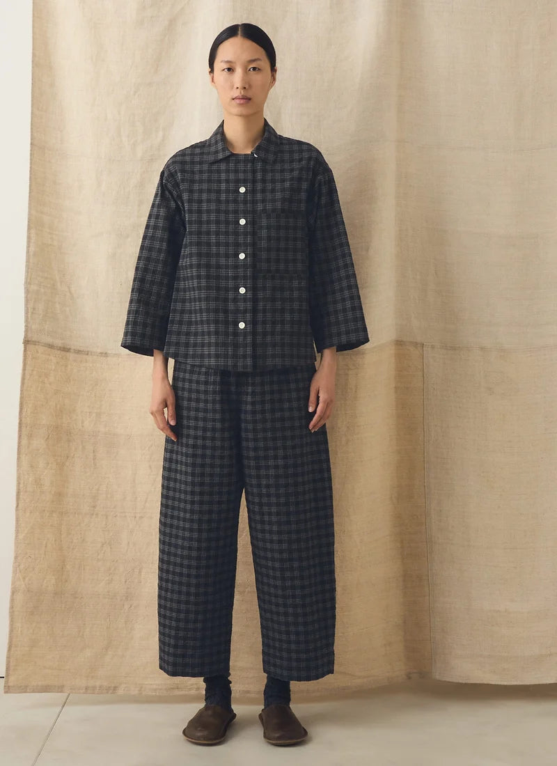 A model stands against a beige linen backdrop wearing a dark gray and black checkered two-piece set consisting of a long-sleeve button-up shirt and matching wide-leg trousers.