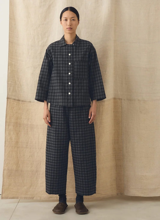 A model stands against a beige linen backdrop wearing a dark gray and black checkered two-piece set consisting of a long-sleeve button-up shirt and matching wide-leg trousers.