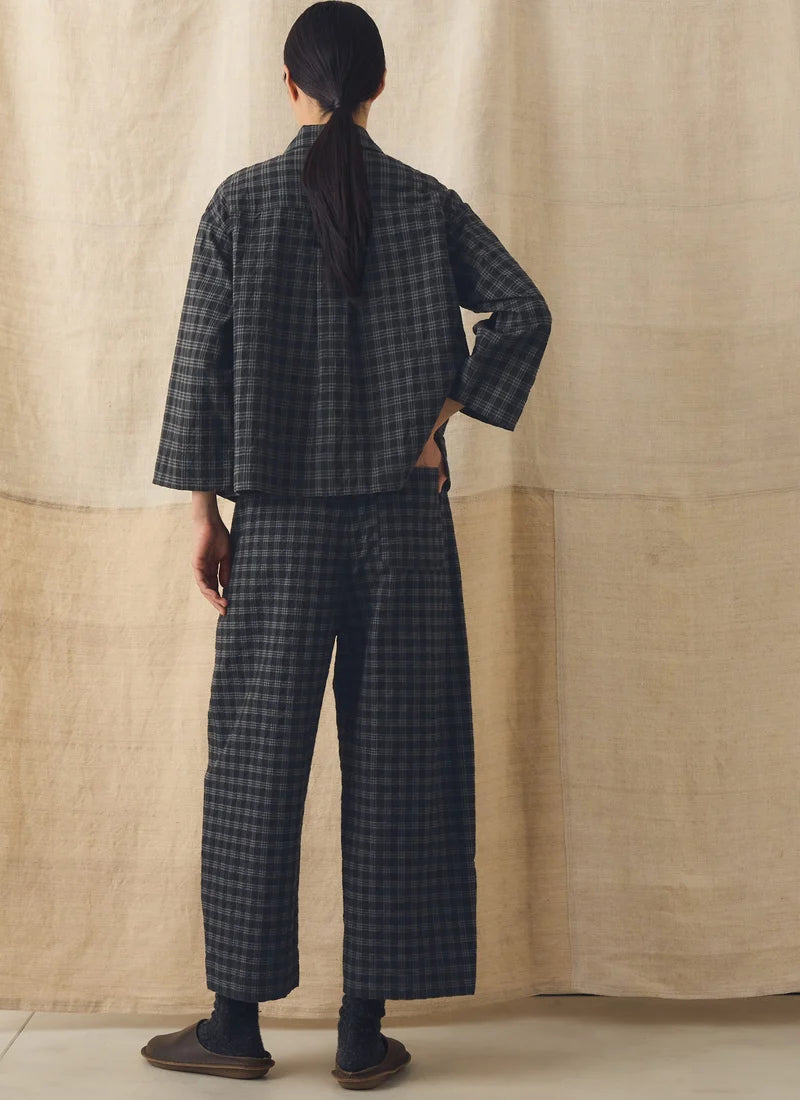 A person shown from the back wearing a dark gray and black plaid two-piece outfit with a cropped jacket and wide-leg pants, standing against a beige fabric backdrop.
