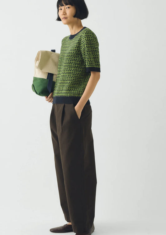 Person wearing a green patterned sweater and brown pants, holding a beige bag on a white background