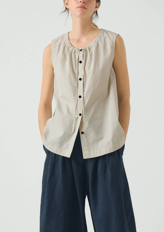 Woman wearing a beige sleeveless button-up shirt and dark pants on a light background