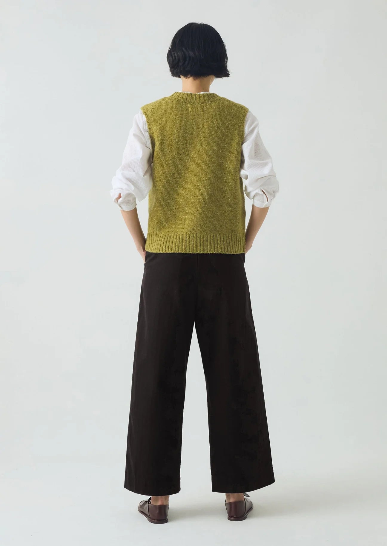 Person wearing a green knitted vest over a white shirt with black pants on a white background