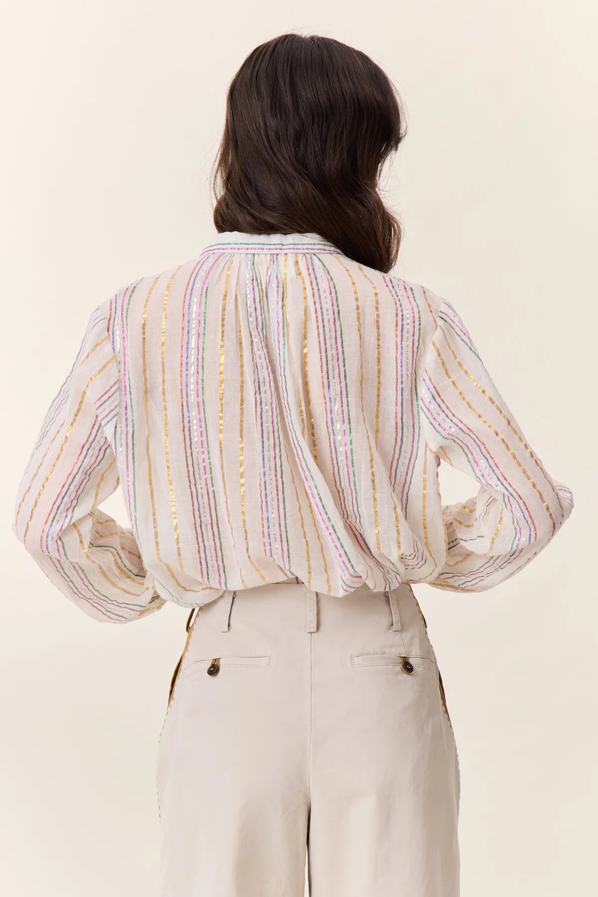 rear view of Woman with long brown hair wearing cream colored pants and cream blouse with colorful metallic stripes