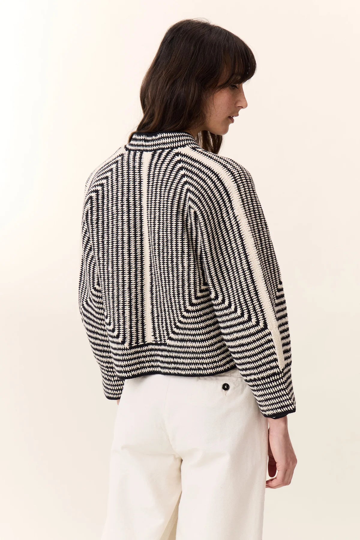 Rear view of photo of woman with long brown hair wearing white pants, black shirt and black and white striped knit cardigan