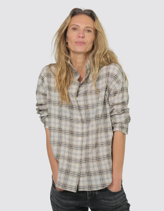 A woman wearing a light beige and gray plaid button-down shirt with dark gray jeans, standing against a solid light gray background.