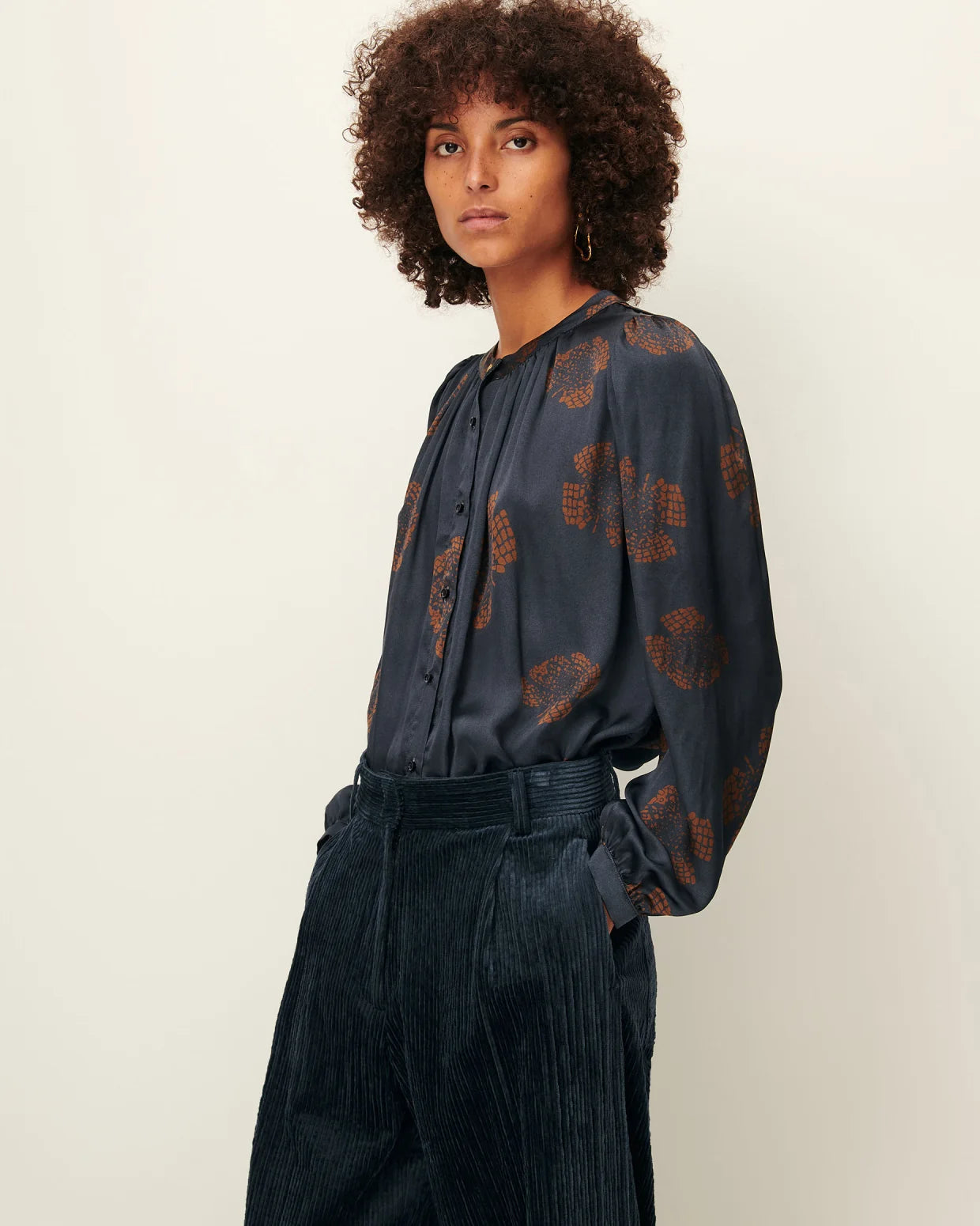 Woman wearing a dark blue blouse with a pattern and dark pants on a plain background