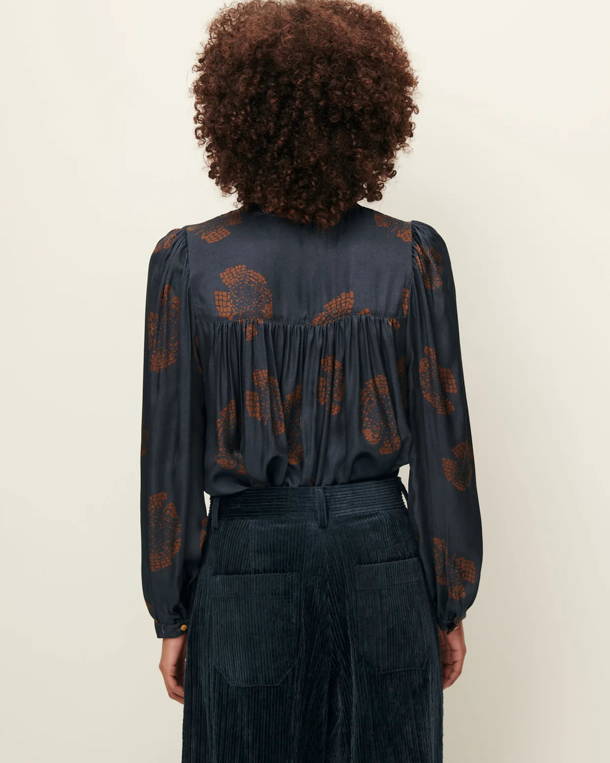 Person wearing a dark blouse with a pattern and dark pants on a plain background